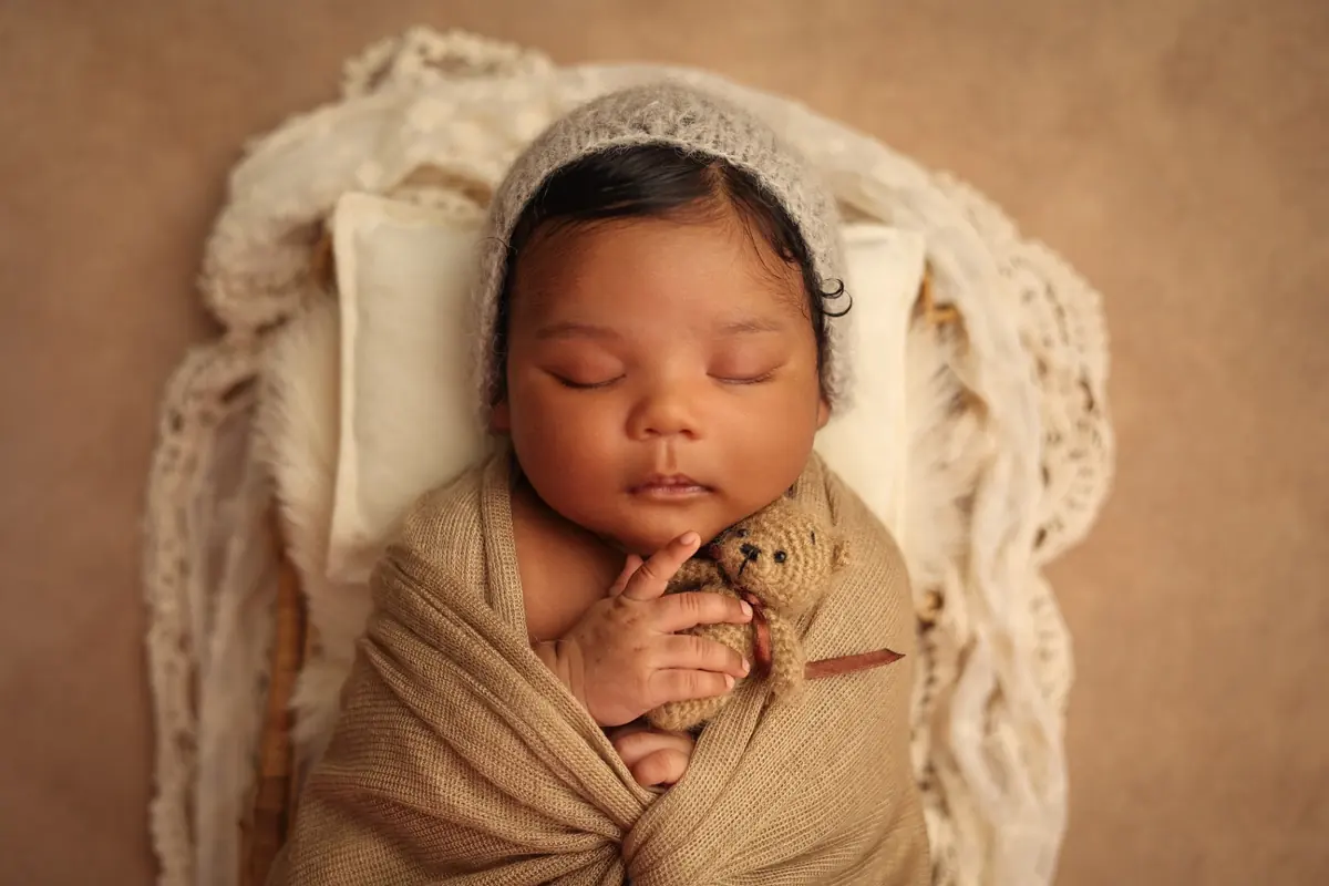 Newborn in a grey knit bonnet cuddling a tiny teddy bear in a basket