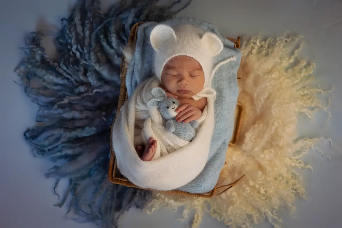Sleeping newborn in a white bear-ear bonnet holding a tiny blue knitted bear in a wicker basket