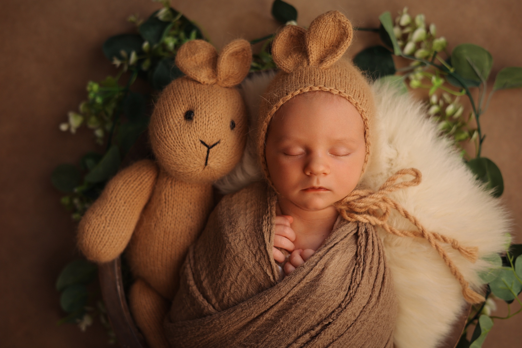 Newborn in a bunny-ear bonnet asleep beside a knitted bunny toy and eucalyptus