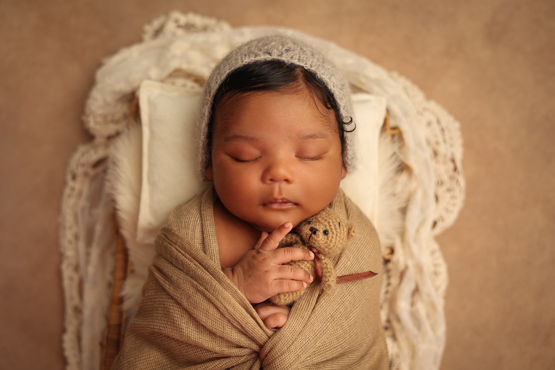 Newborn in a grey knit bonnet cuddling a tiny teddy bear in a basket