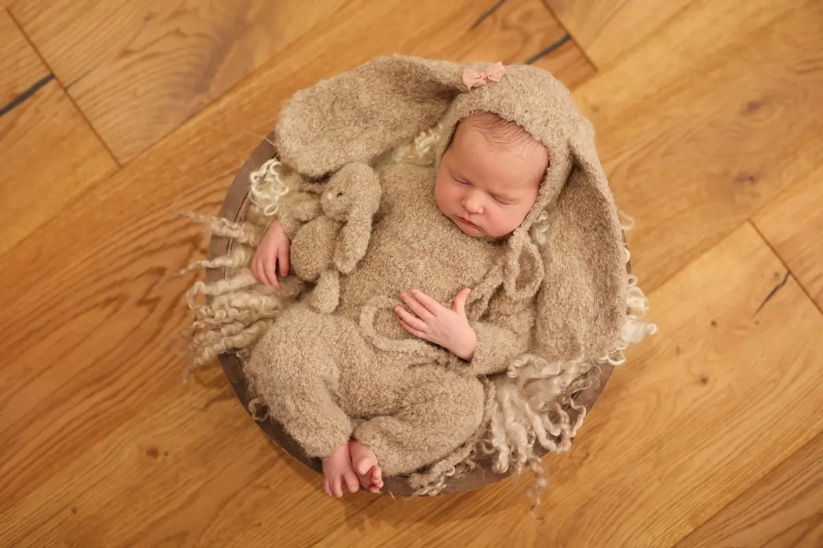 Newborn in a brown bunny-ear knit outfit, asleep in a wooden bowl
