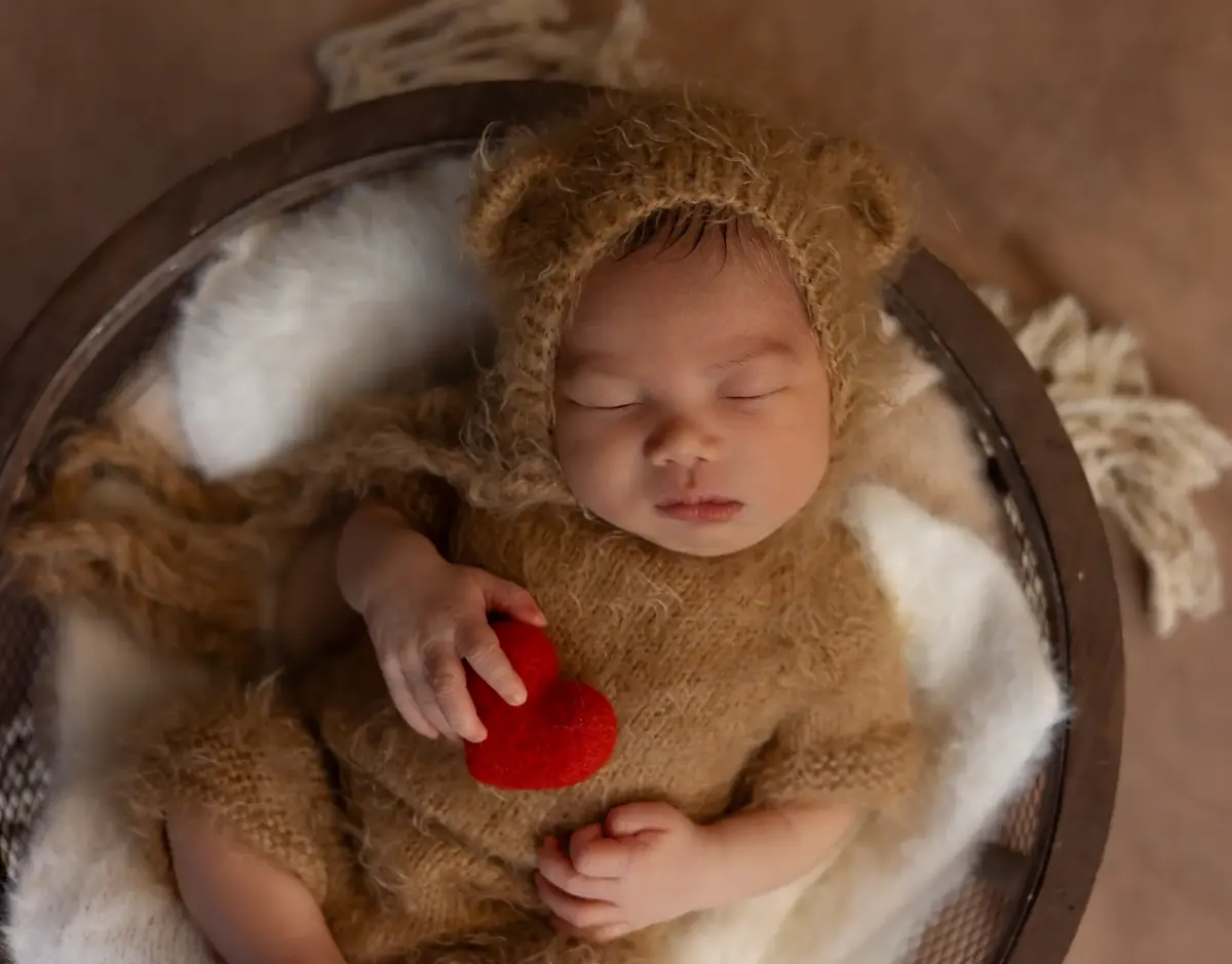 Newborn in a fluffy teddy-bear bonnet holding a tiny red felt heart in a wooden bowl