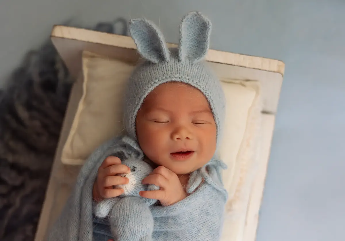 Smiling newborn in a blue knit bunny bonnet cuddling a tiny knitted rabbit in a little wooden bed